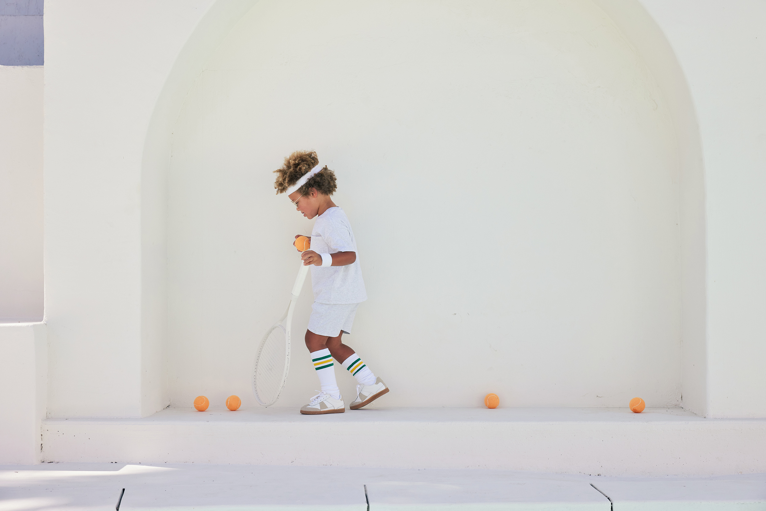 A young child in vintage tennis apparel stands in front of a white wall, holding a tennis racket and ball, with tennis balls on the ground.