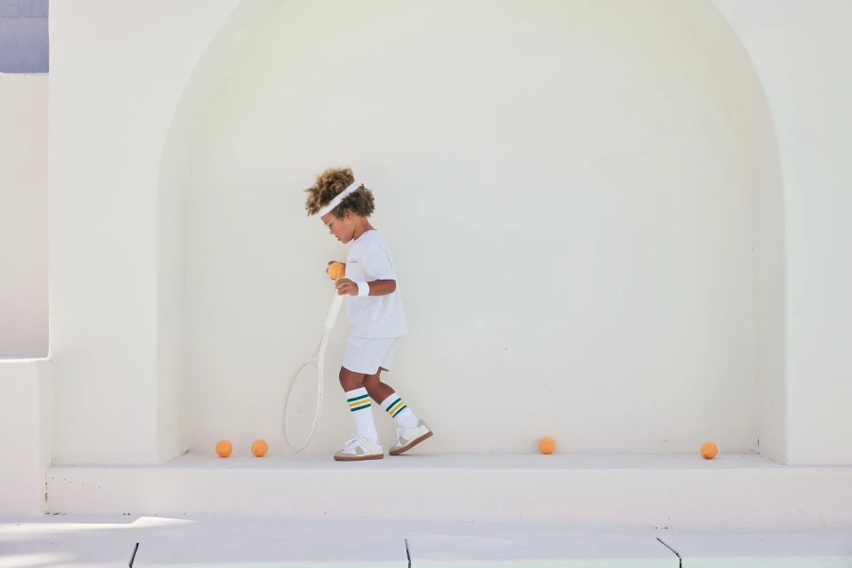 A young child in vintage tennis apparel stands in front of a white wall, holding a tennis racket and ball, with tennis balls on the ground.