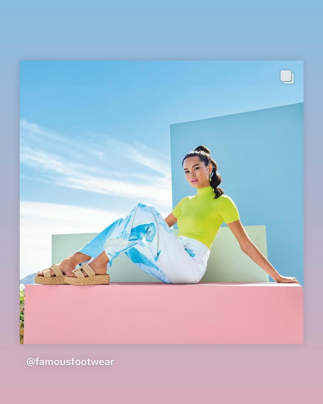 Woman in yellow top and patterned pants sitting in front of blue sky with beige sandals, posing for a fashion or lifestyle ad.