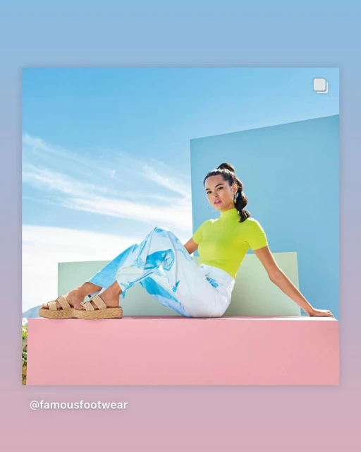 Woman in yellow top and patterned pants sitting in front of blue sky with beige sandals, posing for a fashion or lifestyle ad.