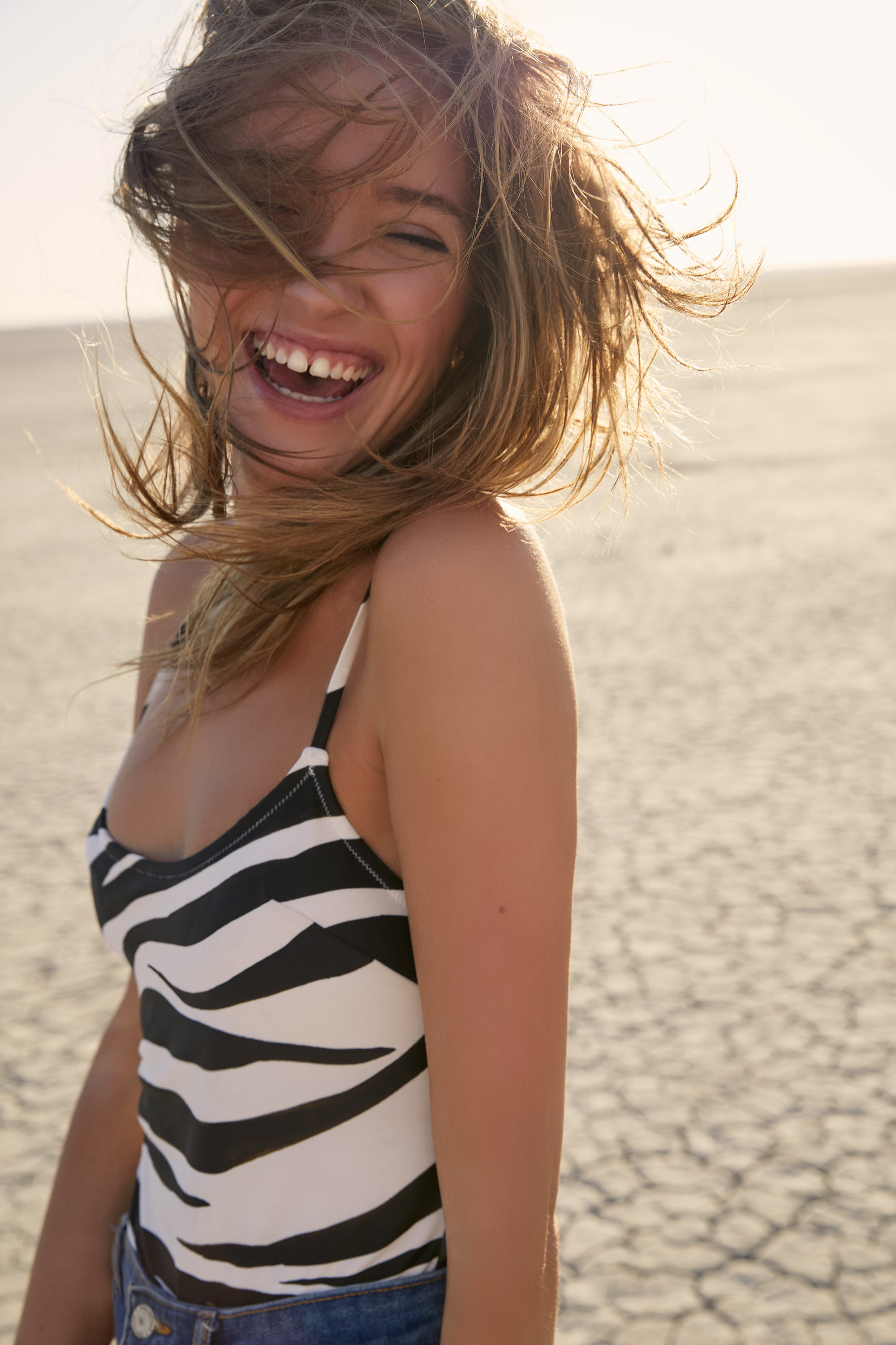 Laughing woman in zebra print swimsuit stands on cracked desert ground with wind blowing hair across face in a sunny, wide shot.
