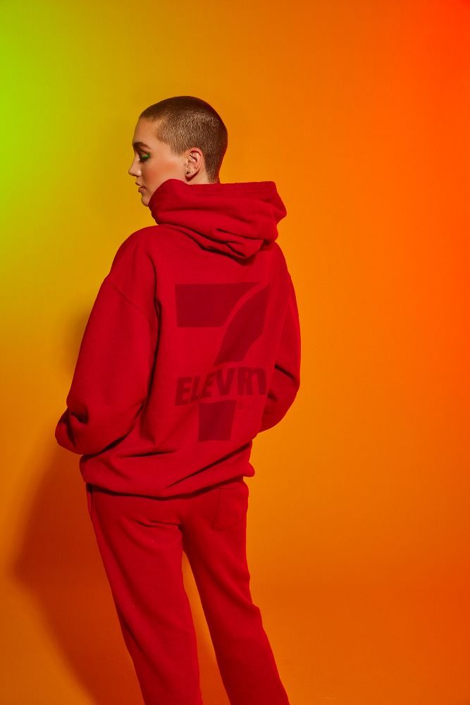 Back view of a young adult woman with short hair wearing a red hoodie and pants, posing in front of an orange and green gradient background.