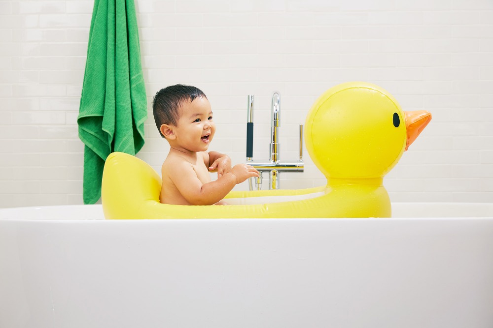 A smiling baby is in a bathtub sitting inside a large yellow inflatable duck in a bright bathroom with a green towel in background.