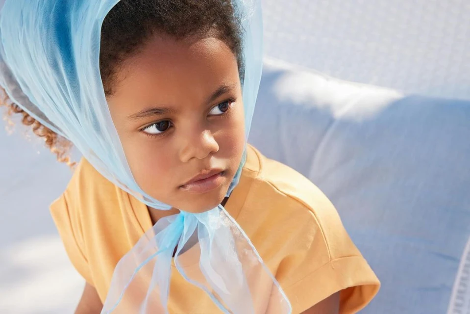 Portrait of a young child with dark hair and skin, wearing an orange top and a sheer blue headscarf, looking to the side.