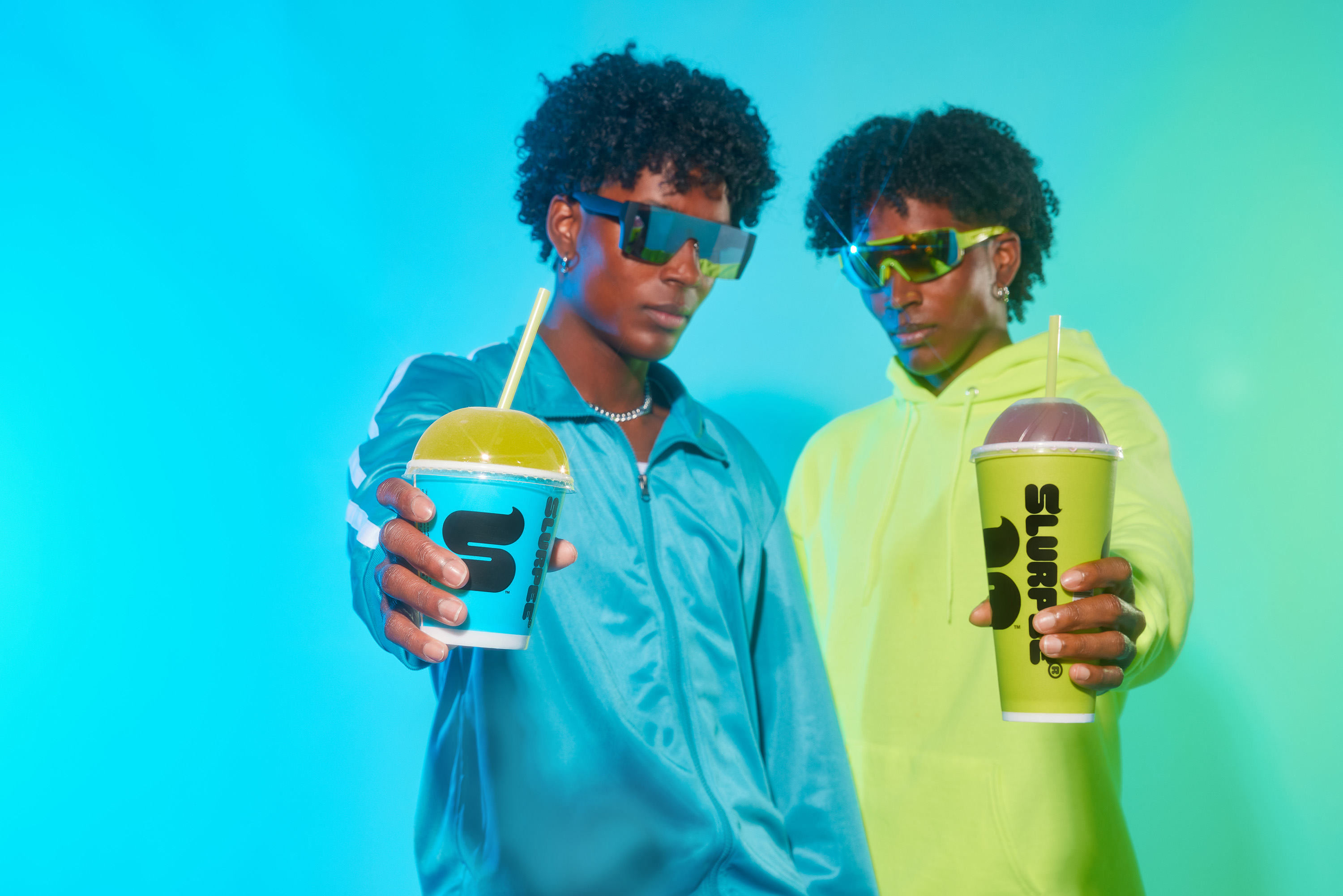 Two young adults pose indoors with blue and green Slurpee drinks, wearing sunglasses and vibrant sporty clothing against a blue background.