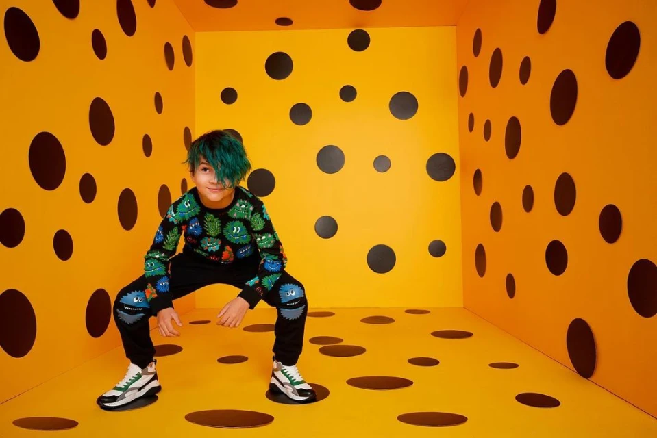 Boy with dyed blue hair wearing a black monster print sweater and pants squats in a yellow room with black polka dots.