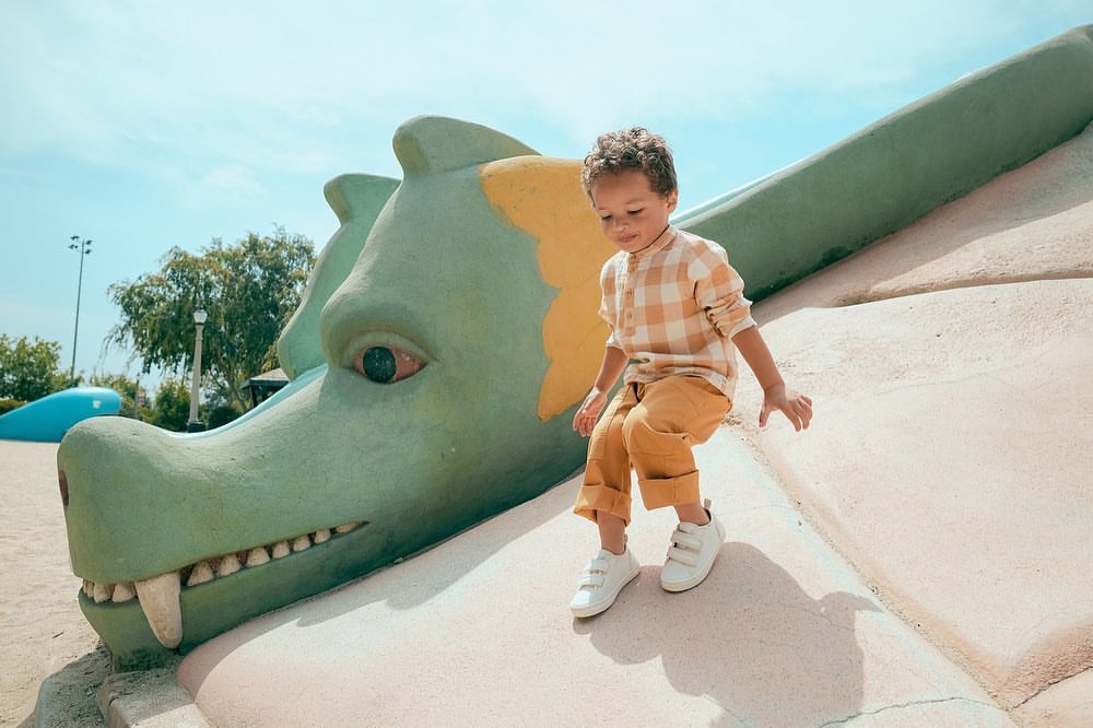 A young child sits on a large dragon sculpture in a park with trees and blue sky. The child is wearing a tan and white plaid shirt.