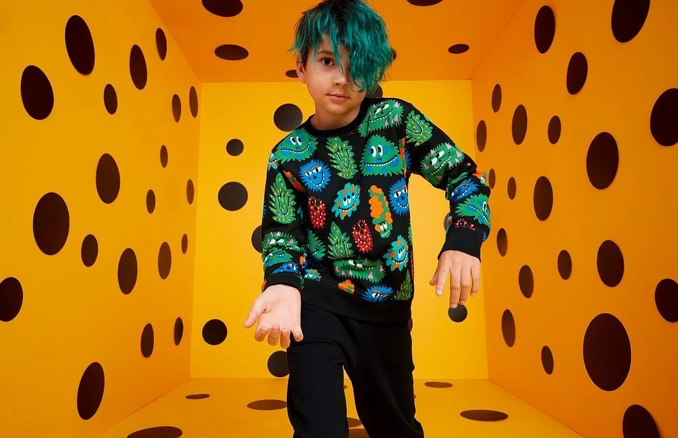 Young boy with teal hair stands in a yellow polka-dot room wearing a monster-themed sweatshirt, captured in a medium shot.