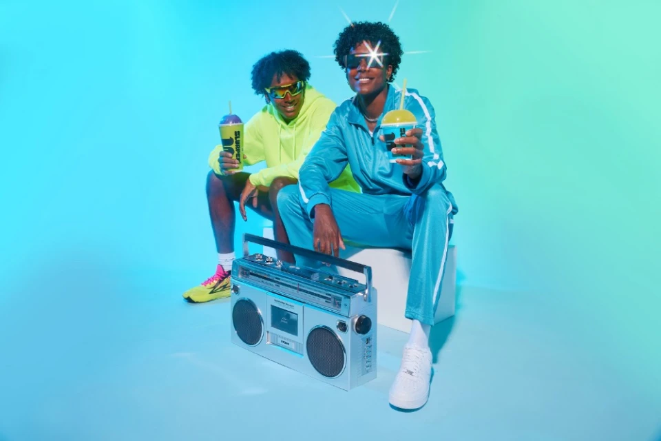 Two adult models pose in a blue photo studio with a retro boombox while holding branded frozen drinks, illuminated by bright, colorful lighting.