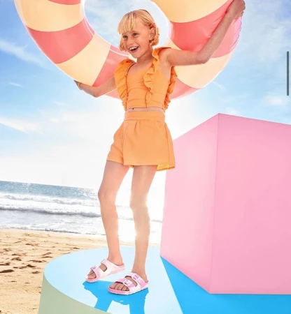 Girl in orange outfit smiles on round platform holding inflatable tube on beach near ocean and oversized pink cube under blue sky.