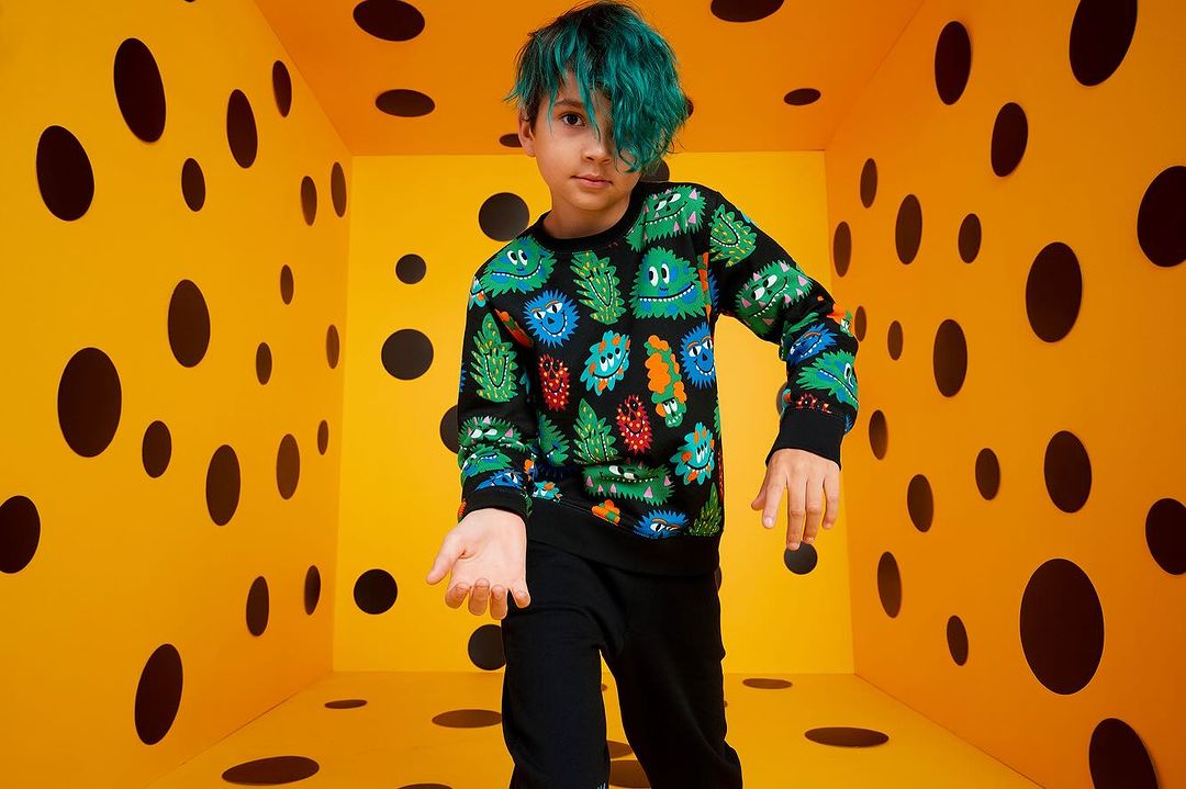 Child model poses in polka-dot room. Young person with blue hair and printed sweatshirt stands in a yellow room with black circles.