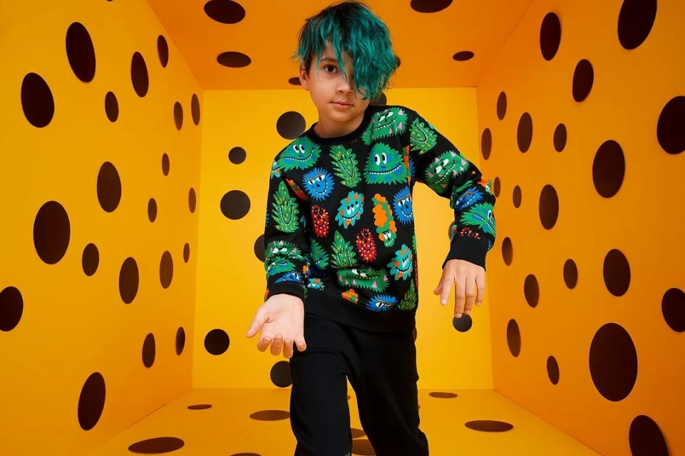Child model poses in polka-dot room. Young person with blue hair and printed sweatshirt stands in a yellow room with black circles.