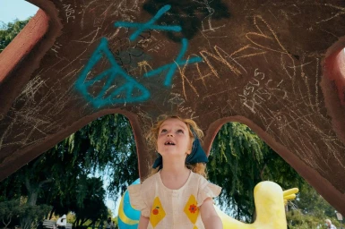 Young child with curly hair wearing a cream patterned dress looking up beneath a painted structure in a park