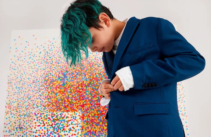 Young person with vibrant teal curly hair fastening a deep blue blazer over a white shirt against a colorful confetti artwork backdrop.