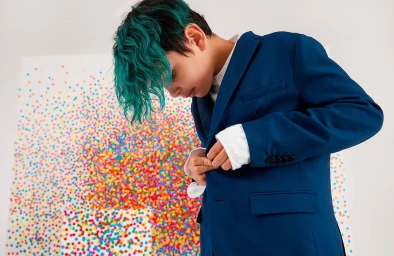 Young person with vibrant teal curly hair fastening a deep blue blazer over a white shirt against a colorful confetti artwork backdrop.