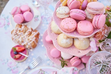 Pink macarons on a tiered dessert stand surrounded by pastries and floral tablecloth