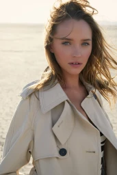 Portrait of a woman with blonde hair wearing a cream trench coat against an outdoor sandy landscape background.