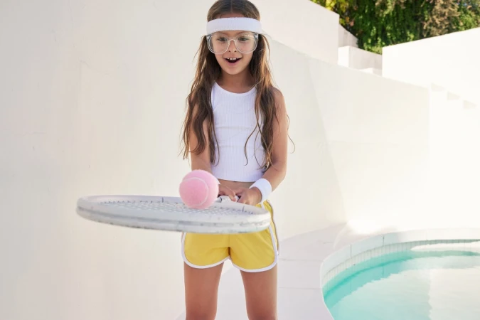 Young child wearing oversized glasses and visor holding a tennis racket near an outdoor pool