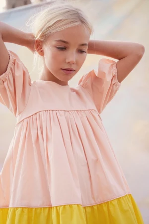Young girl with blonde hair wearing a pink and yellow dress with her hands behind her head against a soft background