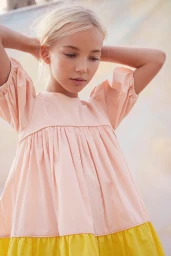 Young girl with blonde hair wearing a pink and yellow dress with her hands behind her head against a soft background