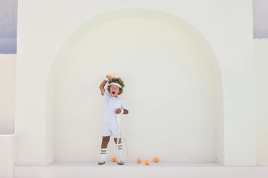 Young child holding a tennis racket and orange balls against a large white arched architectural wall