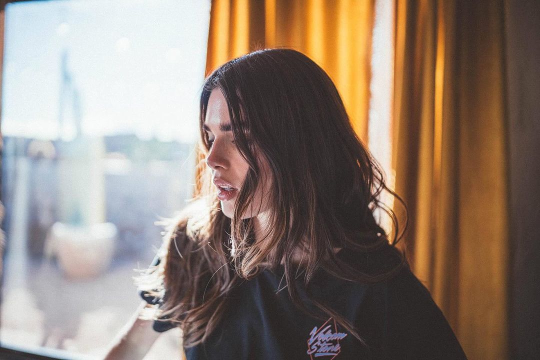 Portrait of an adult woman with long brown hair, wearing a black t-shirt, standing indoors near a window with yellow curtains.