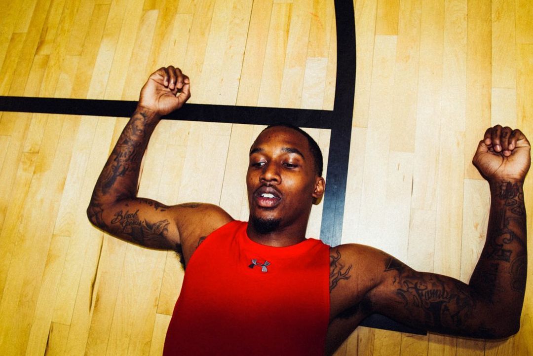 Adult male athlete with tattoos lies on a wood floor in a tank top with arms raised up, with a basketball court line in the background.