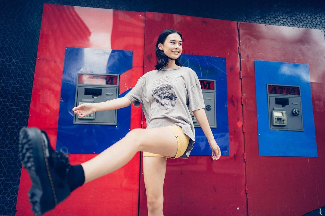 A young woman playfully poses outdoors in front of colorful ATM machines, wearing a graphic tee, yellow shorts, and black boots.