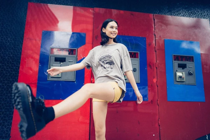 A young woman playfully poses outdoors in front of colorful ATM machines, wearing a graphic tee, yellow shorts, and black boots.