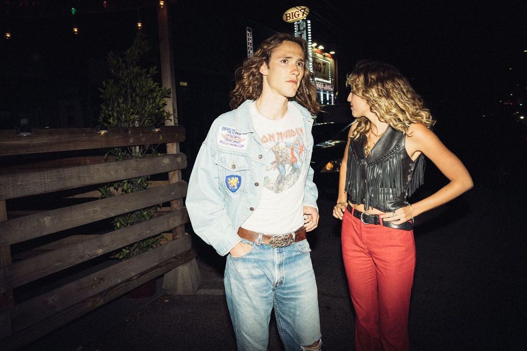 Two young adults wearing retro fashion stand outdoors at night with wooden fence, lights, and signage in the background.
