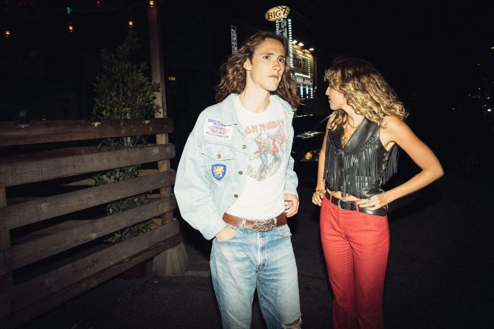 Two young adults wearing retro fashion stand outdoors at night with wooden fence, lights, and signage in the background.