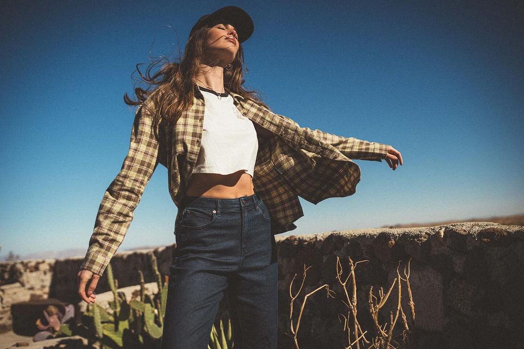 Young woman in a plaid shirt and jeans stands against a bright blue sky with arms outstretched, enjoying the sun outdoors.