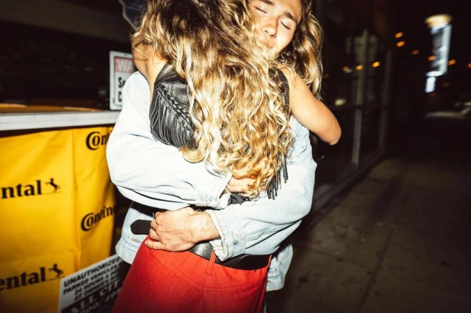 Close-up of a man embracing a woman with curly hair wearing a dark leather top and red pants outdoors at night.