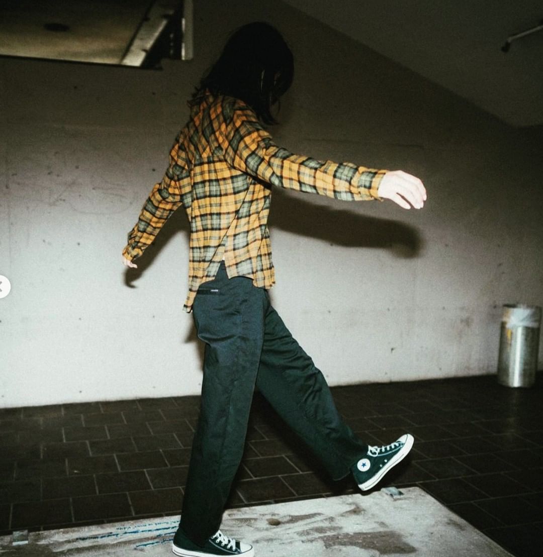 Person in yellow plaid shirt and dark pants walking in an indoor setting with a concrete floor, wearing black sneakers, arms outstretched.
