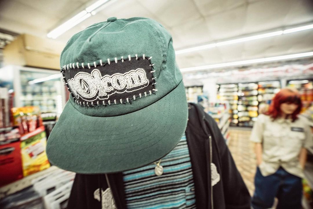A teal baseball cap with a Volcom logo is the central focus, with a woman in a uniform and grocery aisles visible in the background.