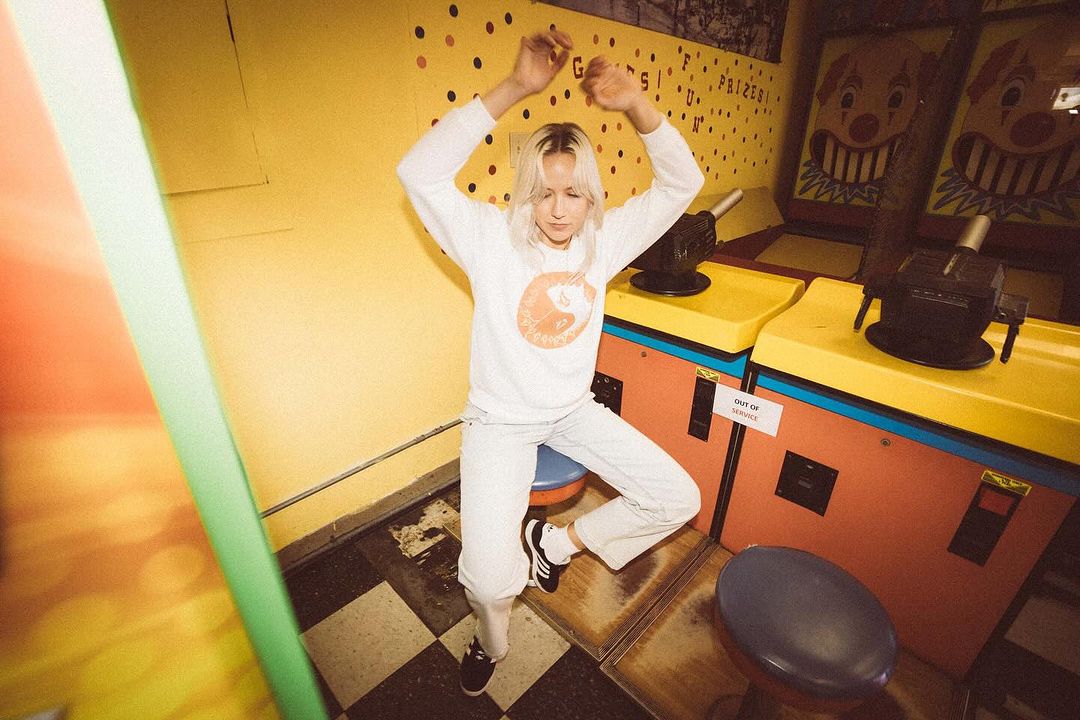 Blonde woman in casual clothes poses with arms raised between vintage arcade machines in a retro-styled game room with checkerboard floor.