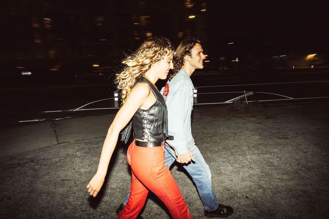 Woman in a leather vest and red pants walks with a man in a light denim jacket and blue jeans on a city street at night.