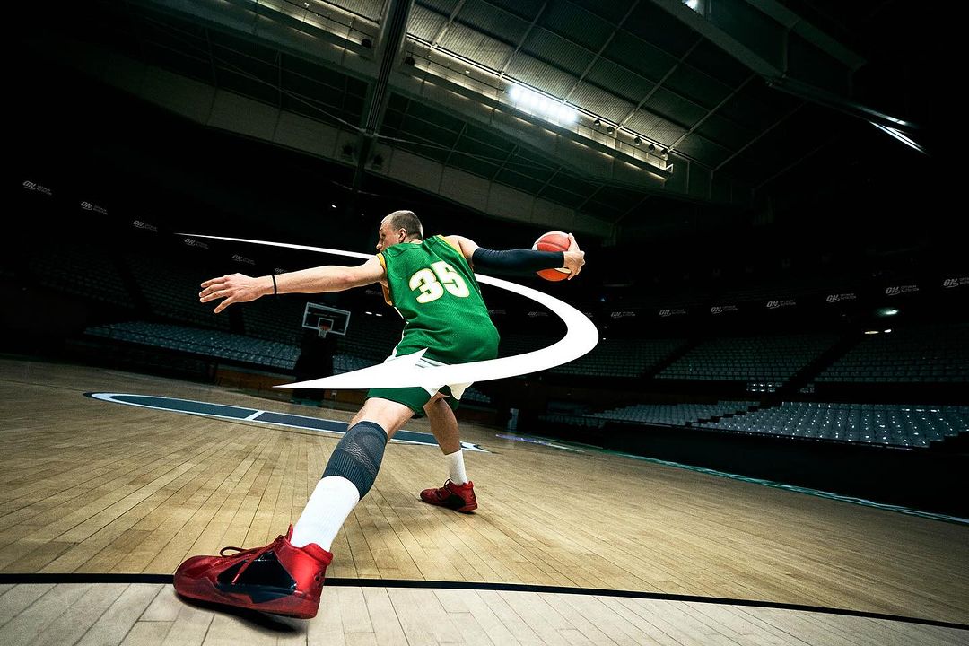 A male basketball player in a green jersey and red shoes shoots a basketball in an indoor court with spectator seating.