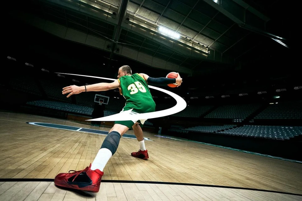 A male basketball player in a green jersey and red shoes shoots a basketball in an indoor court with spectator seating.
