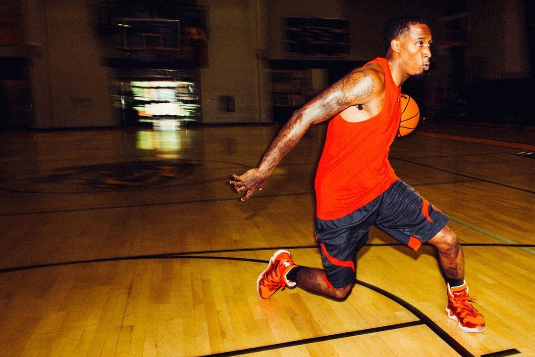 An adult male dribbles a basketball on a hardwood court in a gymnasium, wearing athletic clothes and orange sneakers. The shot is indoors.
