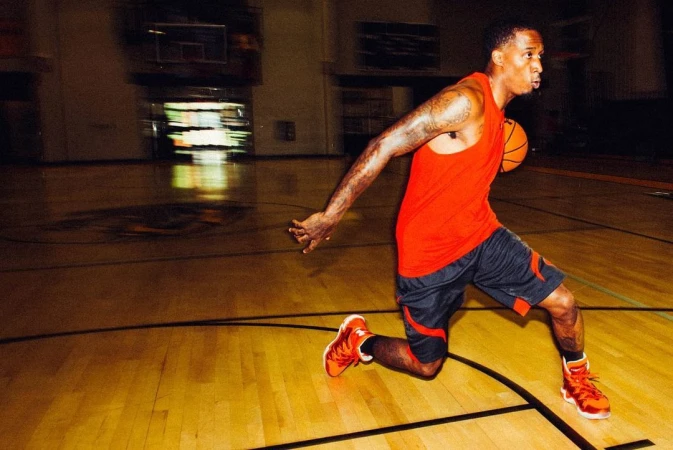 An adult male dribbles a basketball on a hardwood court in a gymnasium, wearing athletic clothes and orange sneakers. The shot is indoors.