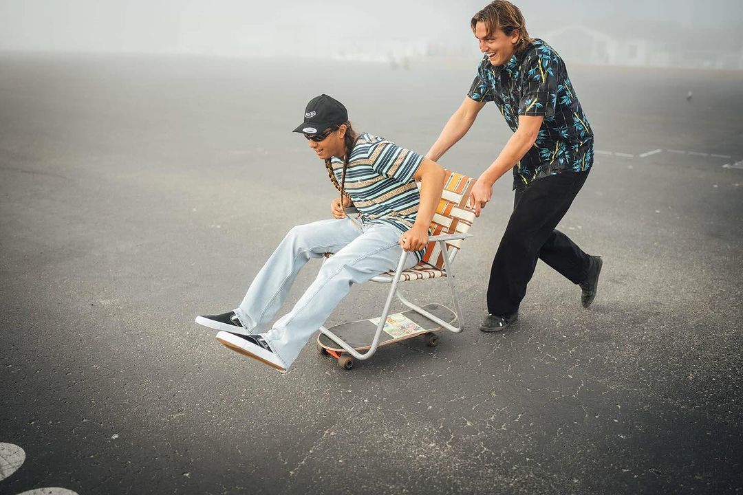 Two young adults share a lighthearted moment with one person pushing the other down a paved road in a chair attached to a skateboard.