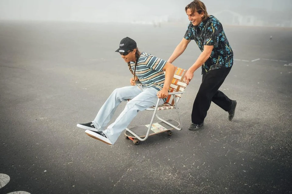 Two young adults share a lighthearted moment with one person pushing the other down a paved road in a chair attached to a skateboard.
