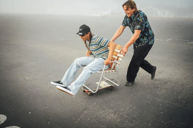 Two young adults share a lighthearted moment with one person pushing the other down a paved road in a chair attached to a skateboard.