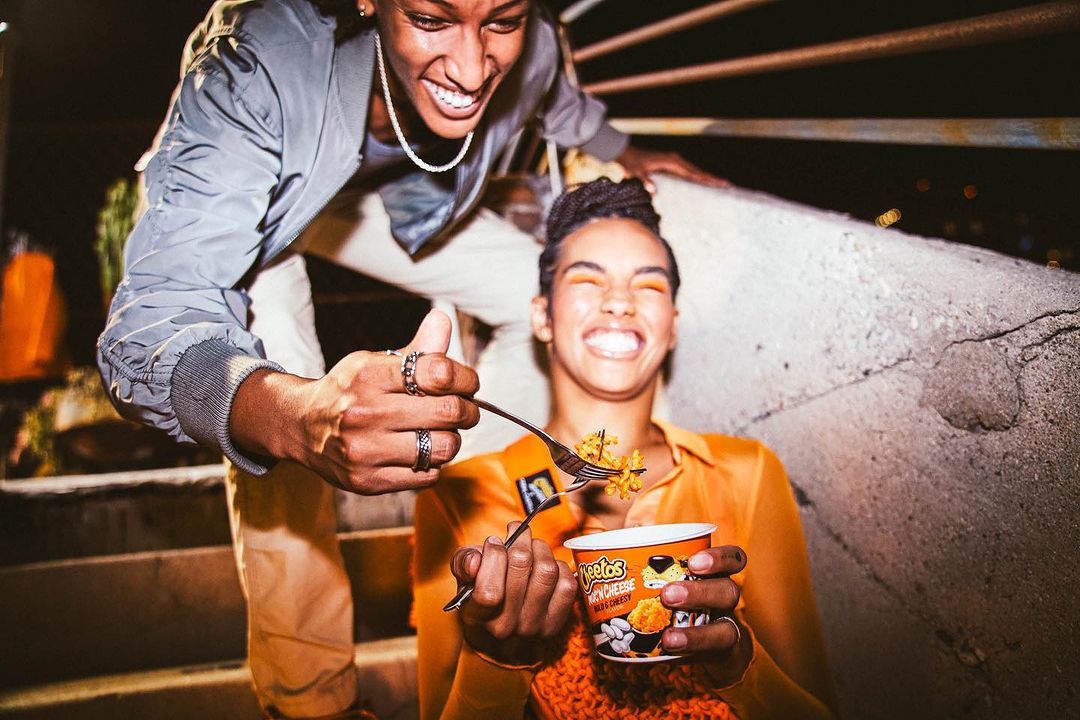 Two adults laugh while one feeds the other a forkful of Cheetos mac and cheese outdoors at night, showcasing product and lifestyle.