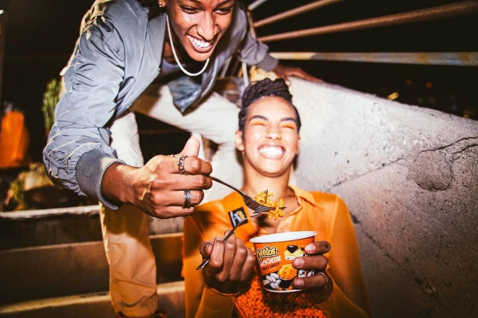 Two adults laugh while one feeds the other a forkful of Cheetos mac and cheese outdoors at night, showcasing product and lifestyle.