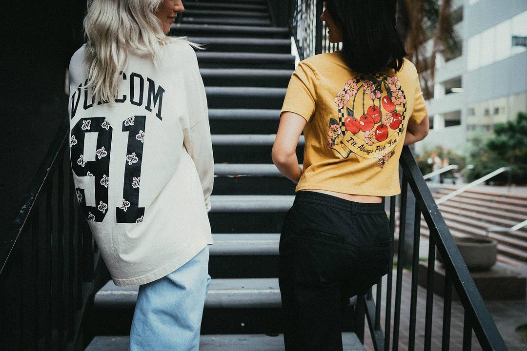 Two women stand on steps. One woman wears a white shirt with 'Volcom 91' on the back. The other wears a yellow shirt with cherries.
