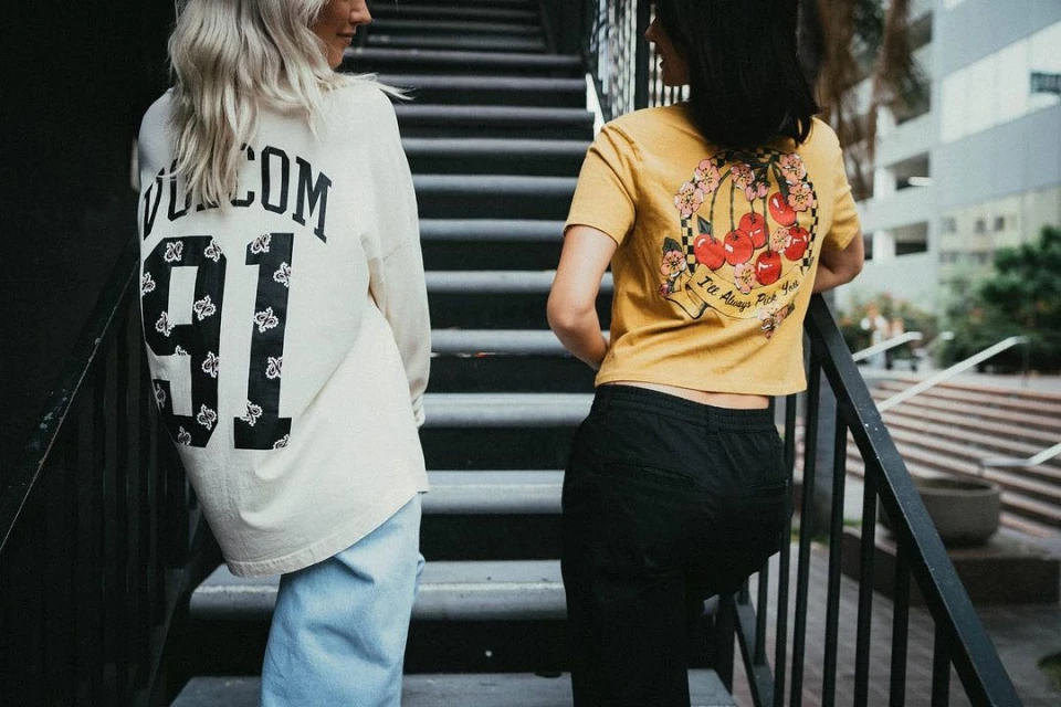 Two women stand on steps. One woman wears a white shirt with 'Volcom 91' on the back. The other wears a yellow shirt with cherries.