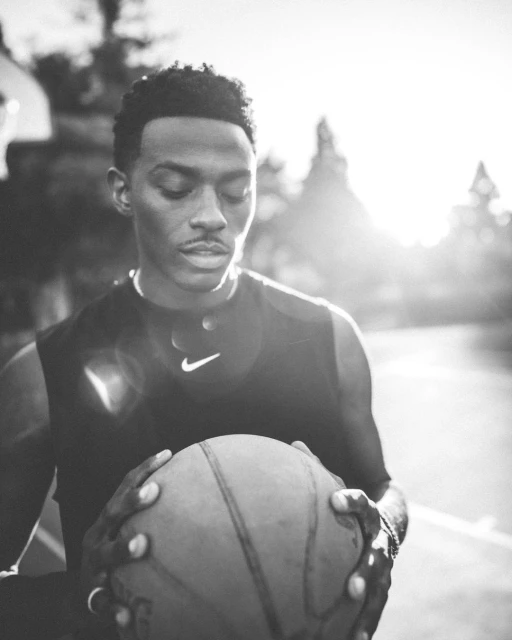 Black and white portrait of an adult male athlete holding a basketball, wearing a Nike tank top outdoors with trees in the background.