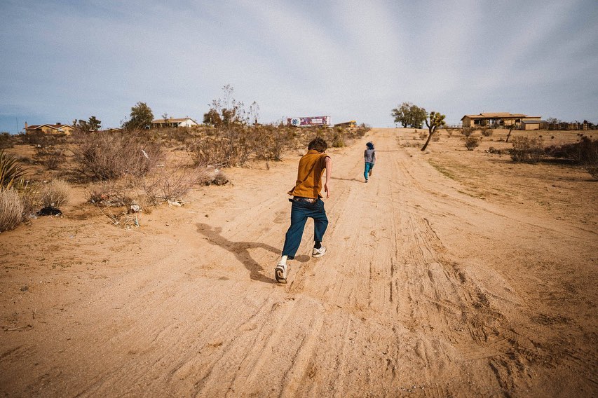 Two people are running along a sandy path in a dry, rural landscape under a blue sky with scattered clouds and low-lying buildings.